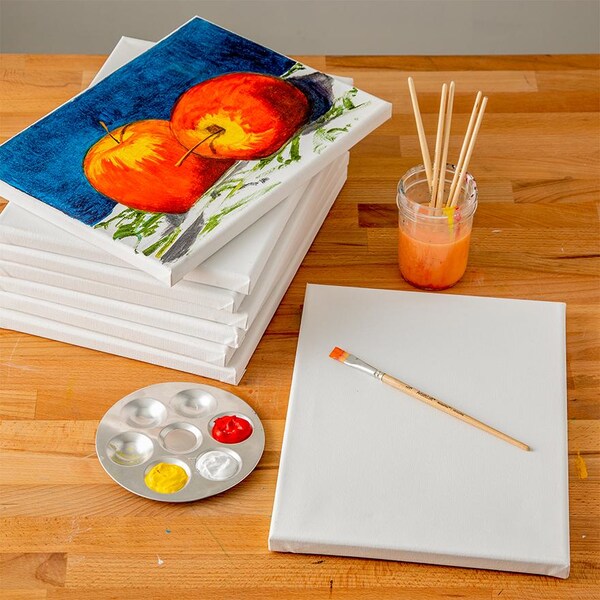 stack of canvas with apple painting next to blank canvas and supplies